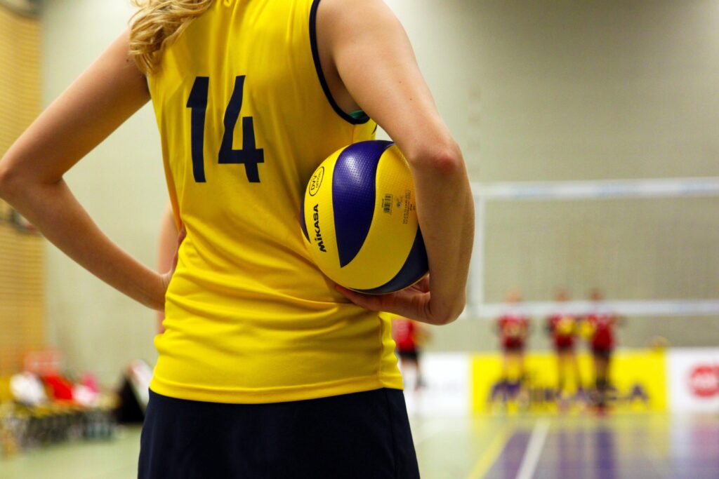 Eine junge Frau in einem gelben Trikot, von hinten fotografiert, die einen Volleyball zwischen ihrem Oberkörper und ihrem Arm einklemmt