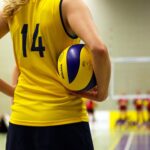 Eine junge Frau in einem gelben Trikot, von hinten fotografiert, die einen Volleyball zwischen ihrem Oberkörper und ihrem Arm einklemmt