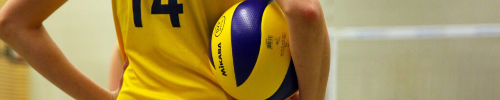 Eine junge Frau in einem gelben Trikot, von hinten fotografiert, die einen Volleyball zwischen ihrem Oberkörper und ihrem Arm einklemmt