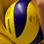 Eine junge Frau in einem gelben Trikot, von hinten fotografiert, die einen Volleyball zwischen ihrem Oberkörper und ihrem Arm einklemmt