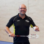 Porträt Andreas Ziegler, junger Mann mit wenig Haaren und Brille, im schwarzen Sportshirt mit Badminton-Schläger in der Hand