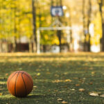 Ein Basketball liegt auf einer Wiese, dazu liegen Blätter auf dem Boden, im Hintergrund unscharf ein Fußballtor und ein Basketballkorb