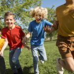 Vier Kindern in bunter Kleidung, zwei Mädchen und zwei Jungs, laufen auf einer Wiese in Richtung der Kamera und lachen dabei