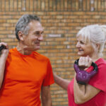 Zwei ältere Personen, eine Mann und eine Frau, mit Kettlebells ihren Händen, vor einer Ziegelwand