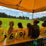 Ein Gruppe junger Mädchen im Fußballtrikot sitzen auf einer Bierbank, dabei eine Fußballtrainerin