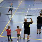 Ein junger Mann zeigt zwei kleinen Jungen wie man Badminton spielt, dazu vielen einige kleine Kinder