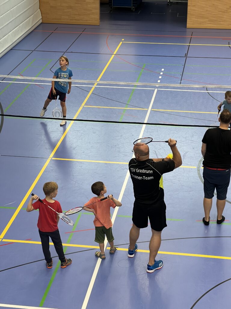 Ein junger Mann zeigt zwei kleinen Jungen wie man Badminton spielt, dazu vielen einige kleine Kinder