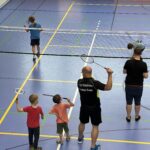Ein junger Mann zeigt zwei kleinen Jungen wie man Badminton spielt, dazu vielen einige kleine Kinder