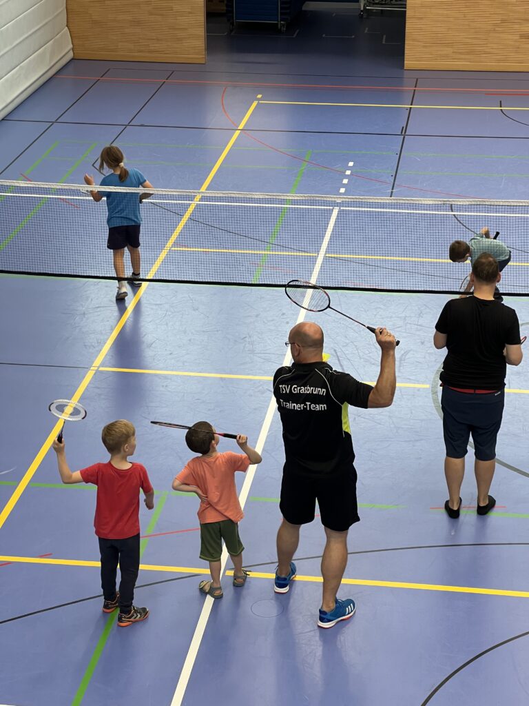 Ein junger Mann zeigt zwei kleinen Jungen wie man Badminton spielt, dazu vielen einige kleine Kinder