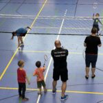 Ein junger Mann zeigt zwei kleinen Jungen wie man Badminton spielt, dazu vielen einige kleine Kinder