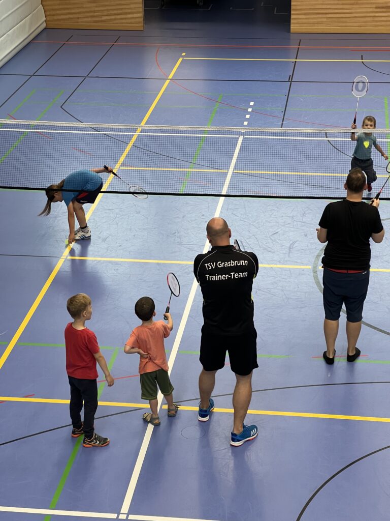 Ein junger Mann zeigt zwei kleinen Jungen wie man Badminton spielt, dazu vielen einige kleine Kinder