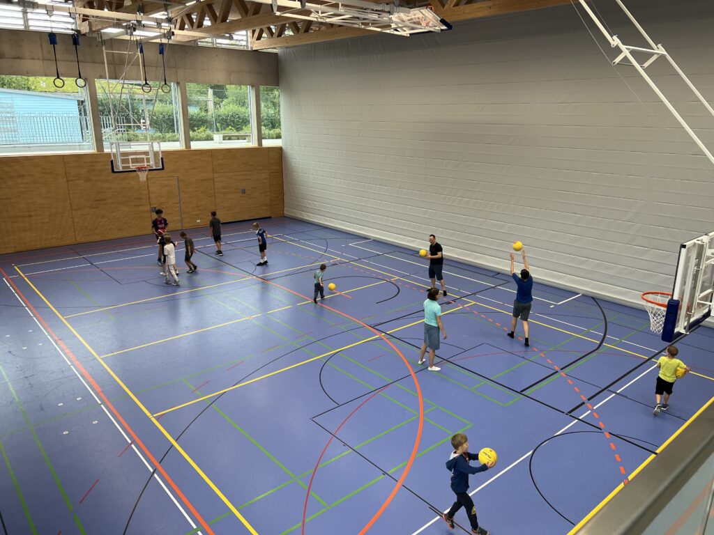 Eine Sporthalle mit vielen kleinen Kindern, die Basketball spielen und dazu einige Eltern