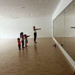 Eine junge Tanzlehrerin in einem Gymnastikraum mit vielen Kindern, die gemeinsam eine Choreografie üben