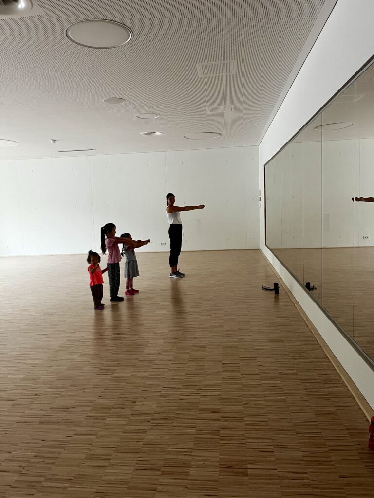 Eine junge Tanzlehrerin in einem Gymnastikraum mit vielen Kindern, die gemeinsam eine Choreografie üben