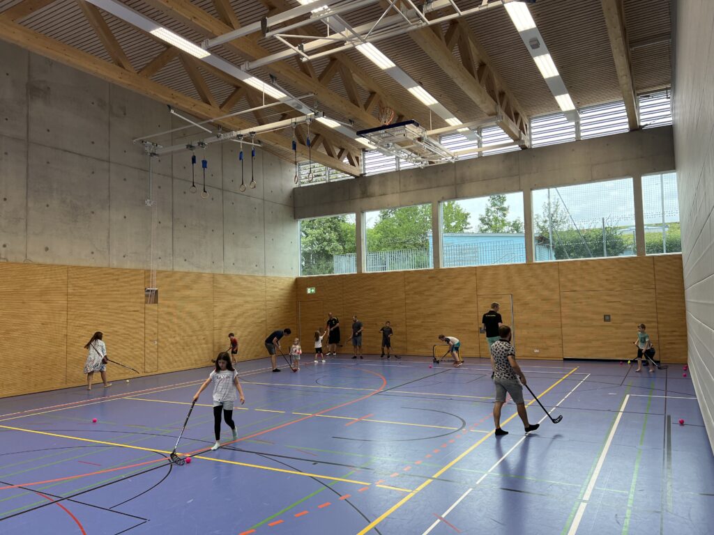 Eine Sporthalle mit vielen kleinen Kindern, die Floorball spielen und dazu einige Eltern