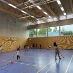 Eine Sporthalle mit vielen kleinen Kindern, die Floorball spielen und dazu einige Eltern