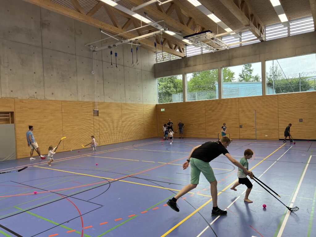 Eine Sporthalle mit vielen kleinen Kindern, die Floorball spielen und dazu einige Eltern