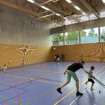 Eine Sporthalle mit vielen kleinen Kindern, die Floorball spielen und dazu einige Eltern