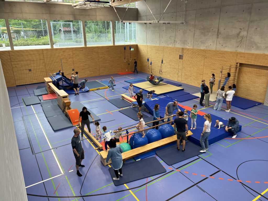 Eine Sporthalle mit einem Hindernisparcour, dazu viele kleine Kinder und einige Eltern