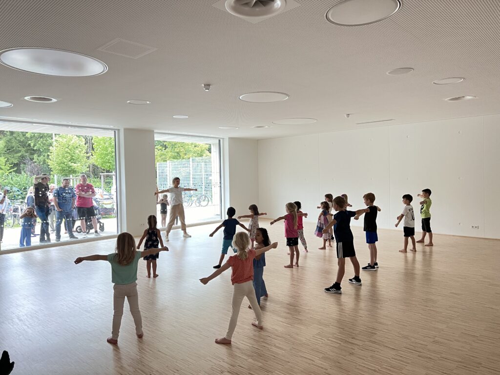 Eine junge Tanzlehrerin in einem Gymnastikraum mit vielen Kindern, die gemeinsam eine Choreografie üben