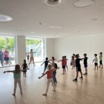 Eine junge Tanzlehrerin in einem Gymnastikraum mit vielen Kindern, die gemeinsam eine Choreografie üben