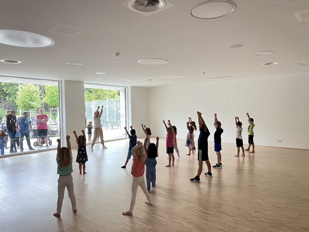 Eine Tanzlehrerin in einem Gymnastikraum mit vielen Kindern, die gemeinsam eine Choreografie üben