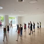 Eine Tanzlehrerin in einem Gymnastikraum mit vielen Kindern, die gemeinsam eine Choreografie üben