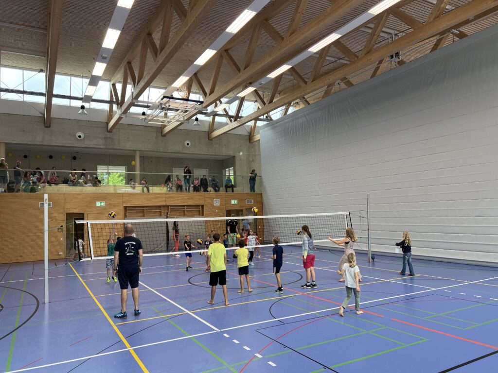 Ein Sporthalle mit einem Volleyball-Netz und vielen Kindern und Jugendliche die Volleyball spielen, dazu Zuschauer auf einer Tribüne