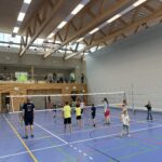 Ein Sporthalle mit einem Volleyball-Netz und vielen Kindern und Jugendliche die Volleyball spielen, dazu Zuschauer auf einer Tribüne