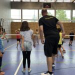 Ein junger Mann zeigt einem kleinen Mädchen wie man Volleyball spielt, dazu vielen kleine Kinder