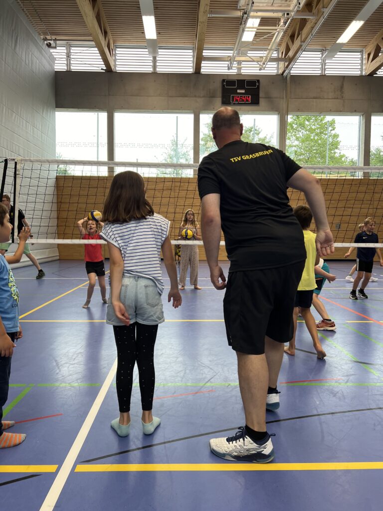 Ein junger Mann zeigt einem kleinen Mädchen wie man Volleyball spielt, dazu vielen kleine Kinder