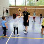Ein junger Mann zeigt einem kleinen Mädchen wie man Volleyball spielt, dazu vielen kleine Kinder