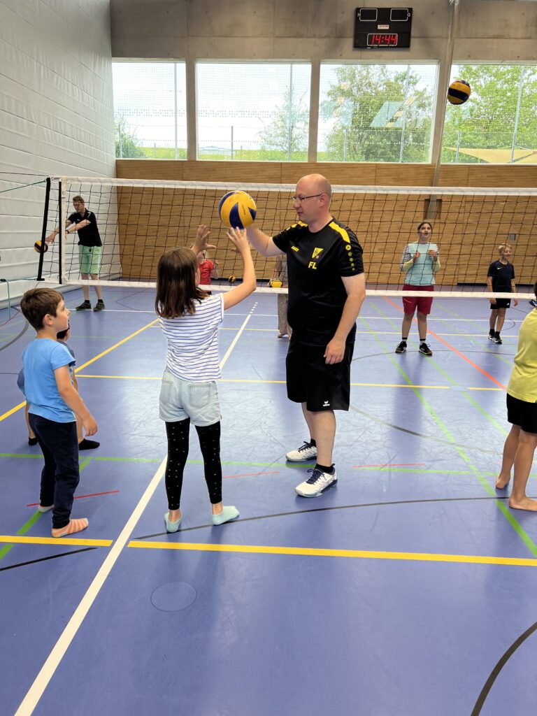 Ein junger Mann zeigt einem kleinen Mädchen wie man Volleyball spielt, dazu vielen kleine Kinder