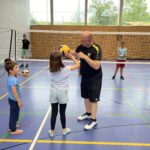 Ein junger Mann zeigt einem kleinen Mädchen wie man Volleyball spielt, dazu vielen kleine Kinder