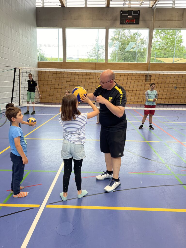 Ein junger Mann zeigt einem kleinen Mädchen wie man Volleyball spielt, dazu vielen kleine Kinder