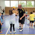 Ein junger Mann zeigt einem kleinen Mädchen wie man Volleyball spielt, dazu vielen kleine Kinder