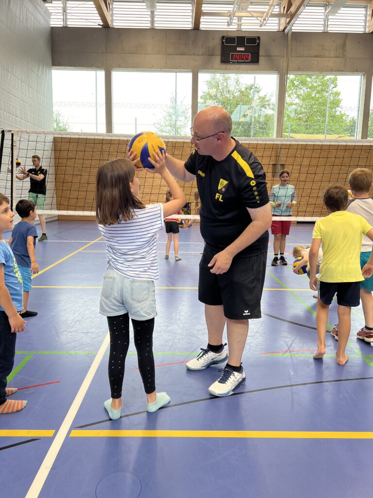 Ein junger Mann zeigt einem kleinen Mädchen wie man Volleyball spielt, dazu vielen kleine Kinder