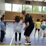 Ein junger Mann zeigt einem kleinen Mädchen wie man Volleyball spielt, dazu vielen kleine Kinder