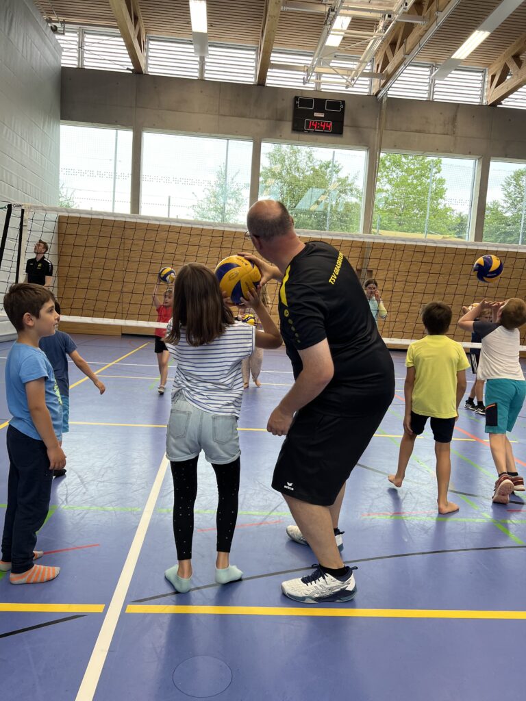 Ein junger Mann zeigt einem kleinen Mädchen wie man Volleyball spielt, dazu vielen kleine Kinder