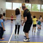 Ein junger Mann zeigt einem kleinen Mädchen wie man Volleyball spielt, dazu vielen kleine Kinder