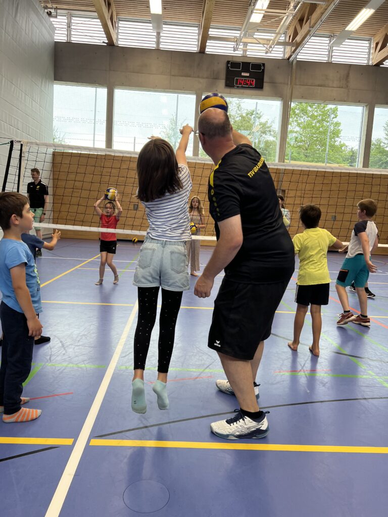 Ein junger Mann zeigt einem kleinen Mädchen wie man Volleyball spielt, dazu vielen kleine Kinder