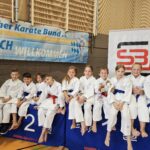 Viele Jungen und Mädchen in Karateanzügen sitzen auf mehreren großen Polstern und schauen in die Kamera