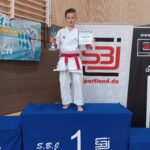 Ein Junge auf einem Podest in einem Karateanzug, der eine Medaille und eine Urkunde in seinen Händen hält