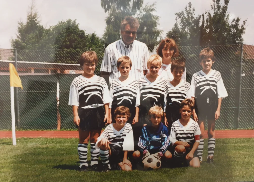 Ein altes Foto von einer einer Fußballmannschaft, dazu ein Trainer, im Hintergrund ein Zaun