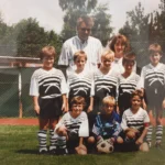 Ein altes Foto von einer einer Fußballmannschaft, dazu ein Trainer, im Hintergrund ein Zaun