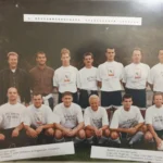 Ein altes Foto von einer einer Fußballmannschaft, dazu mehrere Trainer, im Hintergrund Bäume