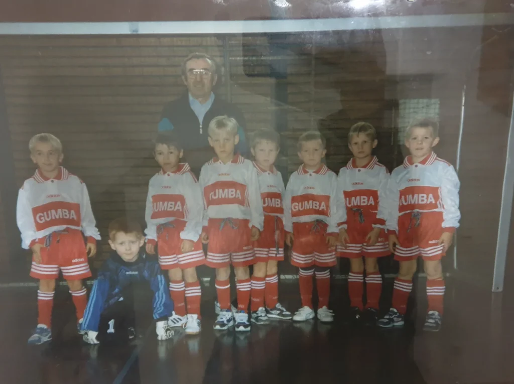 Ein altes Foto von einer einer Fußballmannschaft, dazu ein Trainer, im Hintergrund ein Fußbator