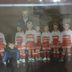 Ein altes Foto von einer einer Fußballmannschaft, dazu ein Trainer, im Hintergrund ein Fußbator