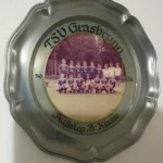 Ein Teller aus Blech, welcher an einer Wände hängt und ein Mannschaftsfoto zeigt, dazu der Text TSV Grasbrunn 1996 Aufstieg B-Klasse