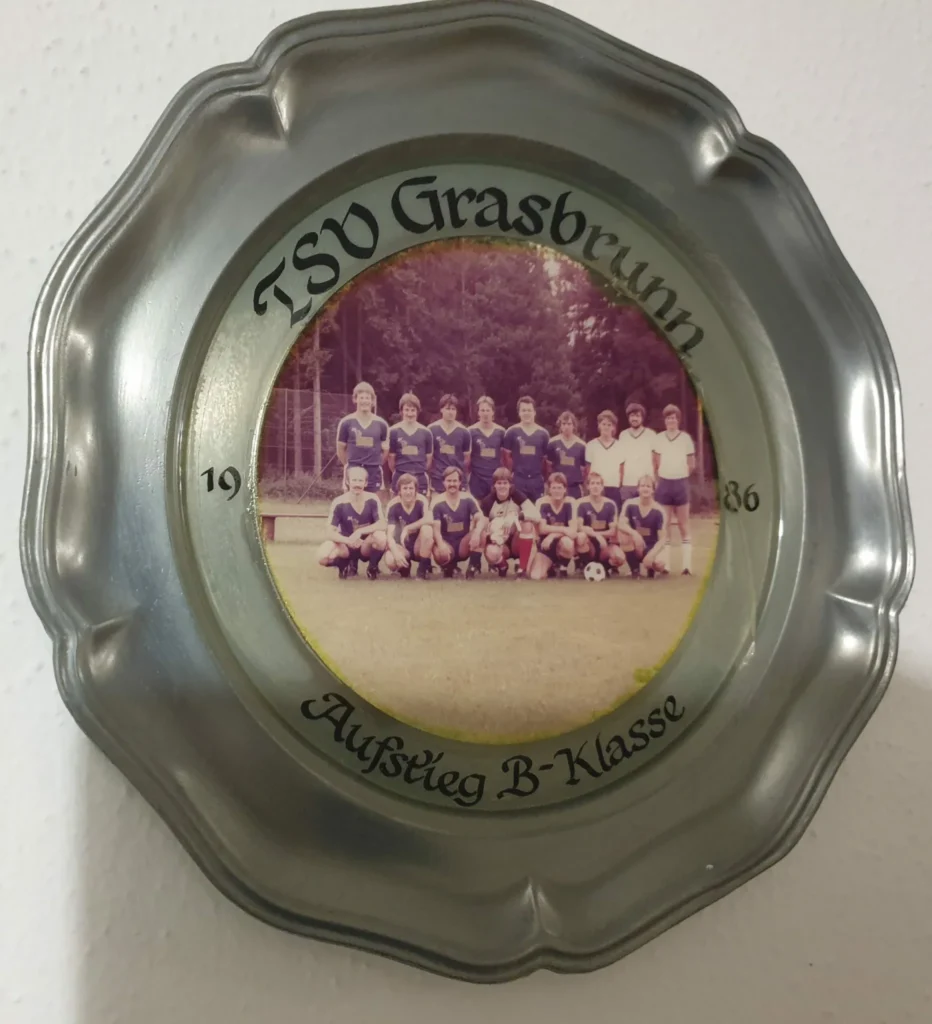 Ein Teller aus Blech, welcher an einer Wände hängt und ein Mannschaftsfoto zeigt, dazu der Text TSV Grasbrunn 1996 Aufstieg B-Klasse